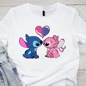 K&ouml;nnte beinhalten: Wei&szlig;es T-Shirt mit einer Grafik von zwei Zeichentrickfiguren, eine blau und eine rosa, die sich mit einem Herzen &uuml;ber ihnen gegen&uuml;berstehen. Das Herz ist zwischen Rosa und Lila geteilt. Die Figuren stammen aus Lilo & Stitch.