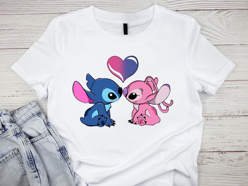 K&ouml;nnte beinhalten: Wei&szlig;es T-Shirt mit einer Grafik von zwei Zeichentrickfiguren, eine blau und eine rosa, die sich mit einem Herzen &uuml;ber ihnen gegen&uuml;berstehen. Das Herz ist zwischen Rosa und Lila geteilt. Die Figuren stammen aus Lilo & Stitch.