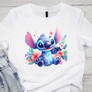 K&ouml;nnte beinhalten: Wei&szlig;es T-Shirt mit einer Aquarell-Illustration der Figur Stitch aus Lilo & Stitch. Stitch ist blau mit gro&szlig;en Ohren und sitzt inmitten von rosa und roten Blumen und gr&uuml;nen Bl&auml;ttern. Das Shirt wird auf einer hellen Holzoberfl&auml;che pr&auml;sentiert.