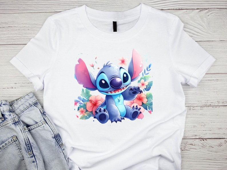 K&ouml;nnte beinhalten: Wei&szlig;es T-Shirt mit einer Aquarell-Illustration der Figur Stitch aus Lilo & Stitch. Stitch ist blau mit gro&szlig;en Ohren und sitzt inmitten von rosa und roten Blumen und gr&uuml;nen Bl&auml;ttern. Das Shirt wird auf einer hellen Holzoberfl&auml;che pr&auml;sentiert.