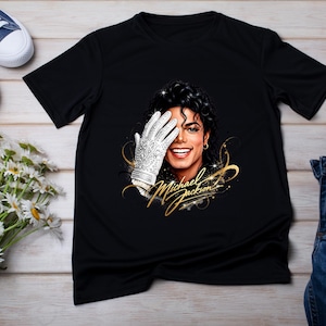 May include: Black t-shirt featuring a portrait of Michael Jackson with a white glove covering his eye. The text "Michael Jackson" is written in gold script. Blue sneakers and jeans are also visible.