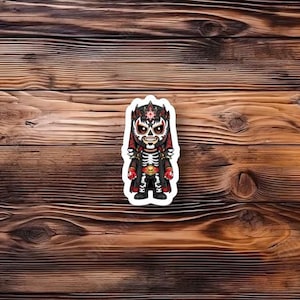 May include: A sticker featuring a stylized illustration of a skeleton character in a luchador mask and outfit. The design includes red, black, and white colors, with intricate details on the mask and clothing. The sticker is placed on a wooden surface.