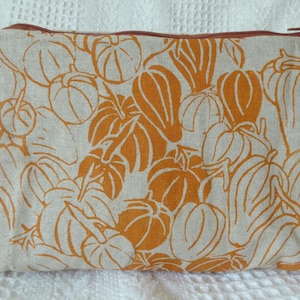 Könnte beinhalten: Eine rechteckige, beige Reißverschlusstasche mit einem sich wiederholenden Muster aus orangefarbenen Kürbissen und Blättern. Der Reißverschluss hat eine dunkelbraune Farbe. Die Tasche ist wahrscheinlich aus Stoff.