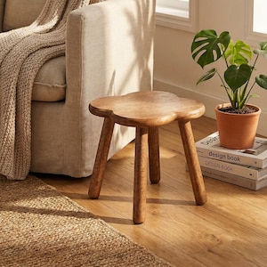 May include: A small, three-legged wooden stool with a clover-shaped top. The stool is made of light brown wood and sits on a hardwood floor. A potted plant and books are nearby, suggesting a cozy interior design.