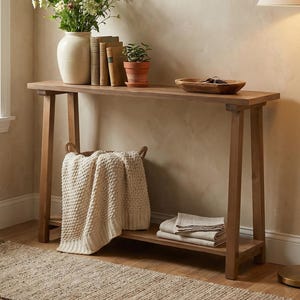 May include: A wooden console table with a lower shelf and a natural finish. The table is adorned with a vase of flowers, books, a small potted plant, a wooden bowl, a woven basket, and a folded blanket. The table is 60 inches long.