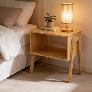 May include: A wooden nightstand with a built-in shelf and legs. A small potted plant and a book sit on top of the nightstand, next to a cylindrical lamp with a light beige shade. The lamp is lit, casting a warm glow.