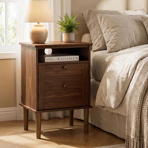 May include: A dark brown wooden nightstand with a lamp, a potted plant, and a book. The nightstand has a drawer and a shelf. The lamp has a beige lampshade. The book on the shelf reads "KINFOLK TABLE".