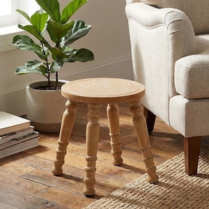 May include: A small, round wooden stool with four turned legs. The stool is light brown and has a simple, classic design. It is placed on a hardwood floor next to a woven rug and a potted plant.