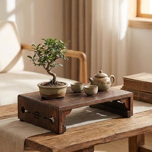 May include: A dark brown wooden tea table with a bonsai tree, teapot, and two small cups. The table is on a larger wooden table with a light-colored runner. A stack of old books is visible.