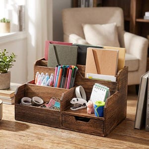 May include: A wooden desk organizer with multiple compartments and a small drawer. It holds pens, notebooks, tape, and a small calendar. The organizer is made of dark wood and sits on a wooden desk. A cup of coffee and a small plant are also on the desk.