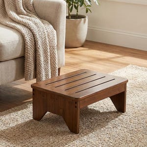 May include: A small, rectangular wooden step stool with a rich brown finish. The top has four evenly spaced slots. The stool is positioned on a woven rug, near a beige armchair and a potted plant.