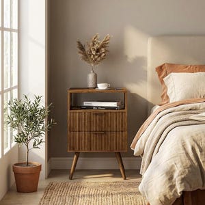 May include: A wooden nightstand with two drawers and a shelf, featuring a ribbed design on the drawer fronts. A vase with dried flowers, a cup, and books are on top. The nightstand is next to a bed with a neutral-toned headboard.