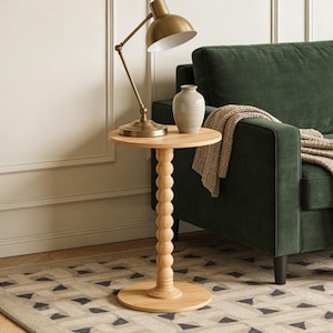 May include: A light wood side table with a round top and a beaded column base. A brass desk lamp and a small vase sit on the table. The table is next to a green sofa. The table is approximately 24 inches tall.