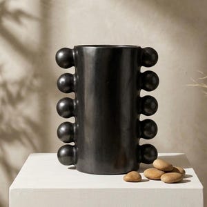 May include: A cylindrical black vase with a smooth finish, featuring a vertical row of five spherical protrusions on both the left and right sides. The vase has an open top and is positioned next to a small cluster of smooth, light brown river stones.