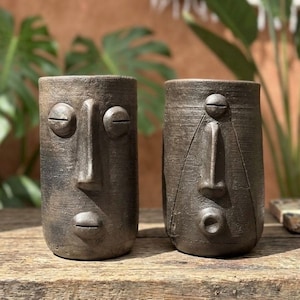 May include: Two cylindrical dark gray ceramic vessels featuring sculpted human-like faces. The left vessel has horizontal slit eyes and a small mouth, while the right vessel features circular eyes and a rounded mouth, both with textured surfaces.