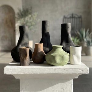 May include: A collection of ceramic vases in various shapes and colors, including black, brown, and green. The vases are displayed on a white, textured pedestal. The background features a neutral-toned wall and natural light.