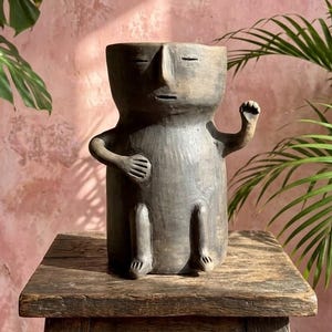 May include: A dark gray, cylindrical ceramic planter shaped like a seated figure with a stylized face, closed eyes, and a small mouth. The figure has one arm resting on its stomach and the other arm raised, featuring a smooth, matte, earthy texture.