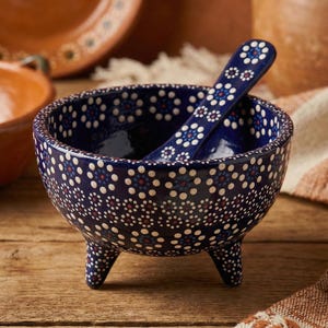 May include: A navy blue ceramic bowl with three legs and a matching spoon. The bowl and spoon are decorated with a pattern of white, red, and blue dots. The bowl is sitting on a wooden surface.