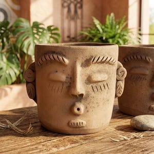 May include: A tan terracotta planter shaped like a human face with closed eyes, arched eyebrows, and a small circular mouth. The surface has a textured, matte finish with subtle dark speckling and carved details on the ears and lips.