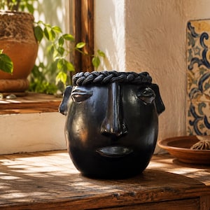 May include: A matte black ceramic planter shaped like a human head, featuring a braided texture along the top rim, a prominent nose, almond-shaped eyes, and defined lips. The vessel has a smooth, dark finish and a rounded base structure.