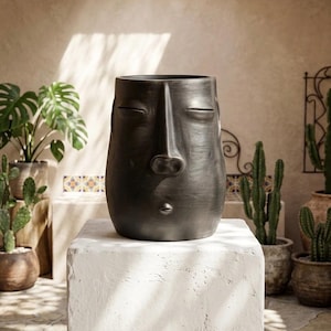 May include: A matte black ceramic planter shaped like a human head with closed eyes, a prominent nose, and small lips. The vessel features a smooth, dark surface and a wide, circular opening at the top, designed to hold plants or decorative items.