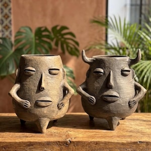 May include: Two anthropomorphic ceramic planters with textured surfaces. The left planter is tan with a smooth face, while the right planter is dark gray with two prominent horns. Both feature closed eyes, a nose, and a mouth, standing on three legs.