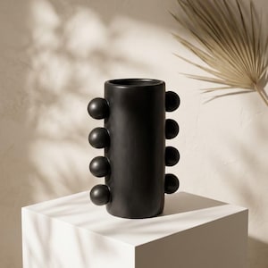 May include: A matte black cylindrical vase featuring a series of four spherical protrusions arranged vertically along each side of the exterior. The vessel has a smooth, uniform finish and a wide, open circular top opening for holding items.