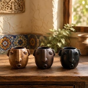 May include: Three ceramic planters shaped like human heads with closed eyes and pursed lips. The planters are arranged in a row, featuring a light brown, a medium brown, and a dark black finish, each with a hollow opening at the top for plants.