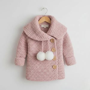 Puede incluir: Un abrigo de ganchillo rosa claro con un cuello grande y botones de madera. El abrigo presenta dos pompones blancos y está colgado de una percha de madera. Las mangas tienen puños doblados.