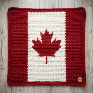 Könnte beinhalten: Ein gehäkeltes Quadrat mit dem Design der kanadischen Flagge. Die Flagge hat einen roten Rand und zwei rote vertikale Balken an den Seiten, mit einer weißen Mitte. Ein rotes Ahornblatt ist auf dem weißen Hintergrund zentriert. Ein kleines Holzabzeichen mit einem Ahornblatt befindet sich in der unteren rechten Ecke.