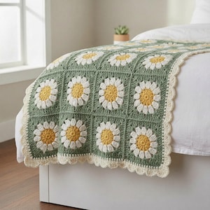 May include: A handmade crochet blanket featuring a daisy pattern. The blanket has a sage green background with white and yellow daisy flowers. The edges are finished with a scalloped cream border. This blanket is perfect for a bed or couch.