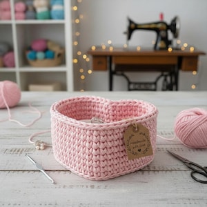May include: A handmade, light pink, heart-shaped crochet basket. A small tag reads "Handmade with Love." The basket sits on a white wooden surface, with a ball of pink yarn, a crochet hook, and scissors nearby. A vintage sewing machine is in the background.