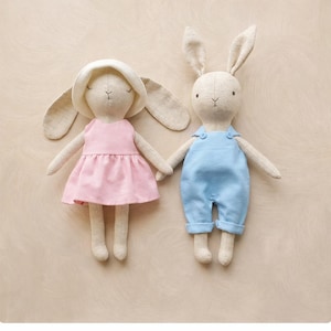 May include: Two handmade bunny dolls. One doll wears a pink dress and a white bonnet. The other doll wears blue overalls. Both dolls are holding hands and have long ears and closed eyes. The dolls are made of a soft, neutral-colored fabric.
