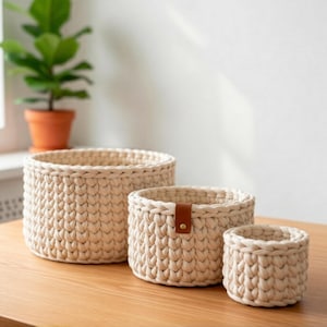 Może przedstawiać: Trzy kremowe, ręcznie robione kosze o różnych rozmiarach. Największy jest otwarty, środkowy kosz ma brązowy skórzany pasek z mosiężnym guzikiem, a najmniejszy to prosty cylinder. Kosze stoją na drewnianej powierzchni.