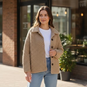 May include: A tan crochet jacket with a collared neckline and button closure. The jacket is worn over a white t-shirt and paired with blue jeans. The jacket has a textured, woven appearance.