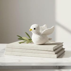 Puede incluir: Una paloma blanca de ganchillo con las alas extendidas, pico amarillo y ojos negros, posada sobre una pila de tres libros de color claro. Una ramita de hojas verdes descansa junto a la paloma. El fondo es de un tono suave y neutro.