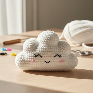 May include: A white crocheted cloud-shaped plush toy with a smiling face, featuring black stitched eyes and a mouth, and pink cheeks. The cloud is on a wooden surface, with crafting supplies in the background.