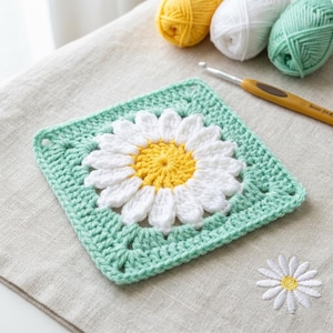 May include: A square, crocheted daisy motif with a yellow center and white petals, set against a light green background. The motif is surrounded by a light green border. Crochet hook and yarn balls in yellow, white, and green are in the background. A small embroidered daisy is also visible.