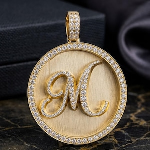 Könnte beinhalten: Goldener Anhänger mit dem Buchstaben "M", besetzt mit kleinen Diamanten. Der Anhänger hat eine runde Form mit einem diamantbesetzten Rand. Der Hintergrund ist goldfarben gebürstet.