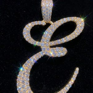 May include: A gold-toned pendant in the shape of the letter "E", encrusted with numerous small, sparkling gemstones. The pendant has a loop at the top for a chain. The gemstones reflect light, creating a brilliant effect against the dark background.