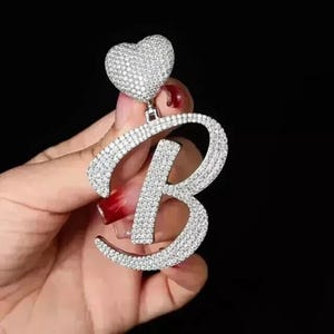 Peut inclure: Un pendentif argenté étincelant en forme de la lettre "B", incrusté de nombreux petits cristaux transparents. Un charm en forme de cœur, également recouvert de cristaux, est attaché au-dessus de la lettre. Le pendentif est sur fond sombre.