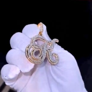 Puede incluir: Un colgante dorado con forma de la letra "R" incrustado con piedras preciosas claras y brillantes. El colgante tiene un lazo en la parte superior para una cadena. El diseño presenta un estilo cursivo con un doble contorno.