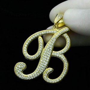 May include: A gold-coloured pendant in the shape of the letter "B", encrusted with numerous small, clear gemstones. The pendant is held against a dark background, showcasing its sparkling details. The jewelry piece is designed with a cursive font.