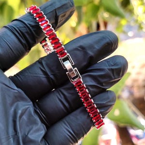 May include: A silver bracelet with rectangular, red gemstones. The bracelet is held by a gloved hand, showcasing the jewelry's design and clasp. The gemstones are closely set, creating a continuous line of color.