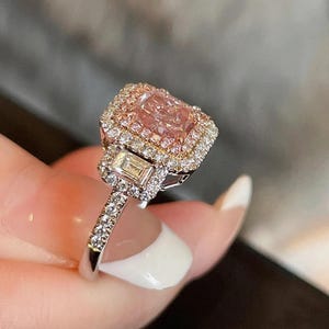 May include: A diamond ring with a large, pink, square-cut central stone, encircled by a halo of smaller diamonds. The silver band is set with more diamonds. The ring is designed to sparkle and catch the light.