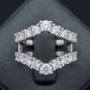 Peut inclure: Une protection de bague en argent avec un design incurvé, sertie de plusieurs pierres précieuses rondes et claires. La bague présente un dessus et un dessous en forme de V, conçus pour s'adapter autour d'une autre bague.