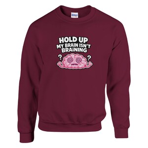 May include: A maroon crew neck sweatshirt featuring the phrase "HOLD UP MY BRAIN ISN'T BRAINING" above a cartoon pink brain with swirling eyes and question marks. The long-sleeved top is a solid colour.