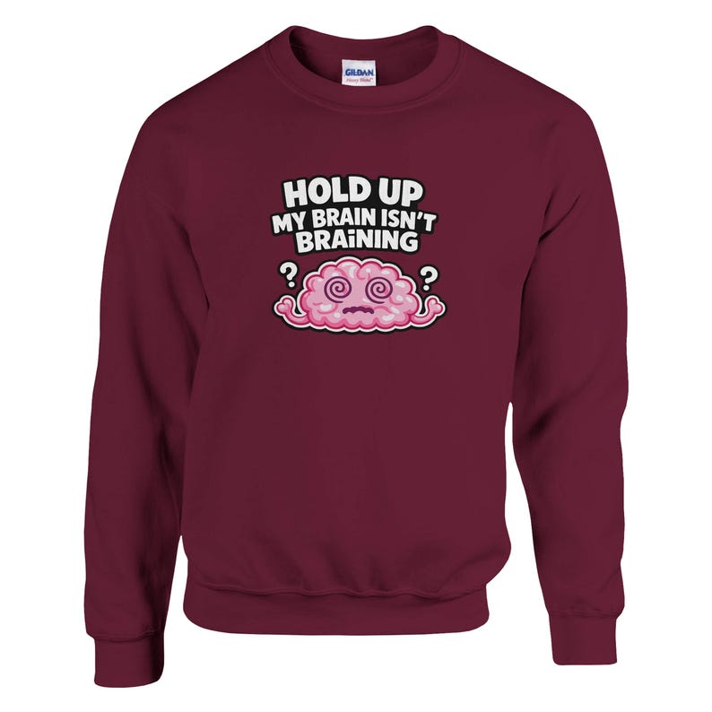 May include: A maroon crew neck sweatshirt featuring the phrase "HOLD UP MY BRAIN ISN'T BRAINING" above a cartoon pink brain with swirling eyes and question marks. The long-sleeved top is a solid colour.