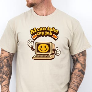 Può includere: T-shirt beige con grafica di computer retrò con una faccina sorridente sullo schermo. Il testo "AI can take my job" è arcuato sopra il computer. Il computer ha le braccia e fa il pollice in su.