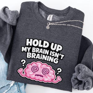 May include: A dark grey sweatshirt featuring the text "HOLD UP MY BRAIN ISN'T BRAINING" in white and black. A pink cartoon brain with swirly eyes and question marks is below the text. A silver necklace with a small pendant is on the sweatshirt.